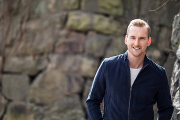 An attractive man standing leaning against a wall of stone wearing a blue jacket and white shirt, feeling relaxed and confident on a sunny day.