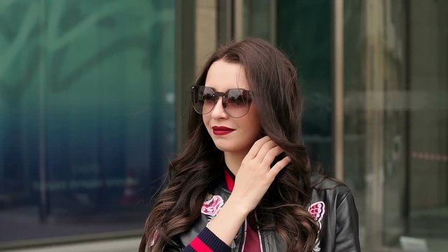 Brunette in sunglasses and a leather jacket stands on the background of a modern building. Portrait of modern girl in sunglasses in the city.