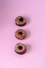 Three baked chocolate doughnuts with chocolate glaze with confetti over pink background. Overhead view.