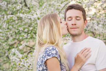 Fototapeta premium Lovely, fallen in love couple at the flowers of tree branches in spring. Young woman kissing a man on cheek.