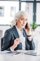 Serious senior businesswoman holding coffee cup and talking on smartphone