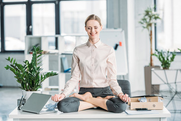 happy businesswoman meditating in lotus position on messy table in office