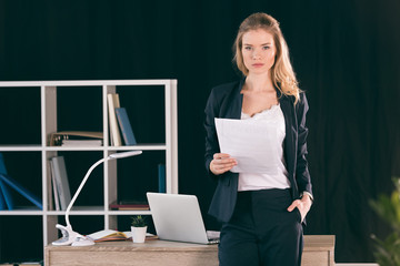 Confident young businesswoman with hand in pocket holding paper and looking at camera