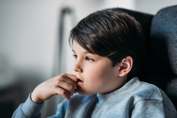 portrait of little adorable pensive boy sitting in armchair and looking away at home