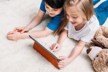 little boy and girl playing with digital tablet while lying on carpet at home