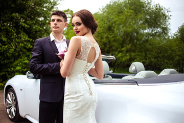 gorgeous bride with fashion makeup and hairstyle in a luxury wedding dress with handsome groom near white cabriolet car