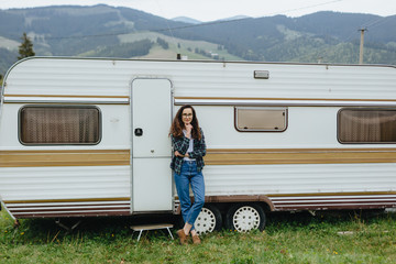 Beautiful girl standing near trailer traveling in mountains. Woman in glasses traveling by car. Traveler.