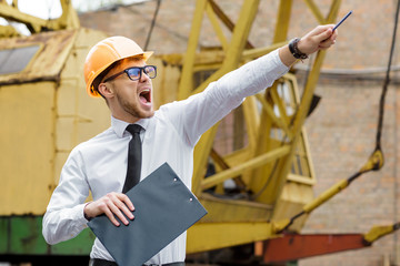 Engineer builder in a helmet holds drawings and shows by hand at construction site