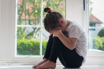 Sad little girl sitting on the window sill hiding her eyes, selective focus