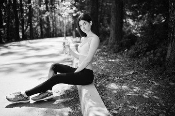 Fitness sport girl in sportswear sitting at road in park with water at bottle, outdoor sports, urban style.