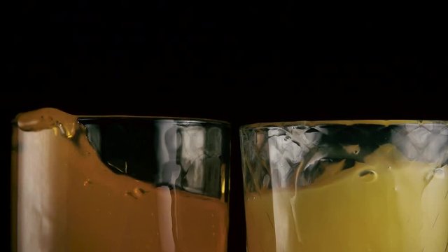 Slow mo. In two glasses with orange drinks falls ice cubes, juice and soda on a black background
