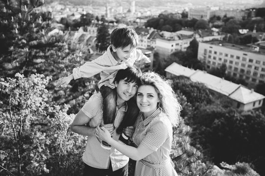 Black and white portrait happy family of three. Father hugging mother and holding his son on the back. They standing and smiling on the hill with city view.