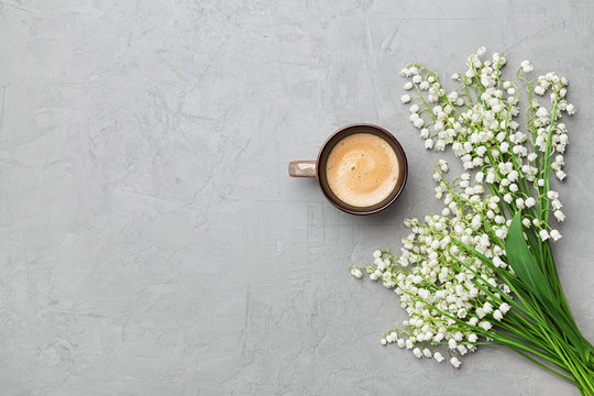 Coffee mug with bouquet of flowers lily of the valley on gray stone table top view in flat lay and minimalistic style. Beautiful morning breakfast.