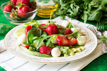 Fresh vegetarian salad with spinach, arugula, avocado slices, strawberries and mini mozzarella on green wooden table. Selective focus