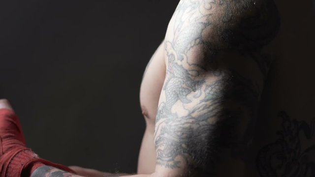 Muscular Muay Thai boxer with tattoos pulling bandage on his hand, slowmotion