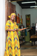 Smiling black African woman in traditional clothes wishing welcome to customers in front of her store
