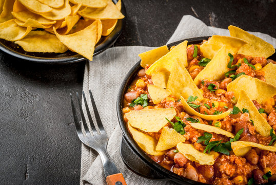 Traditional Mexican Chilean food, chili con carne, served in a portioned skillet pan with nachos tortilla. On black concrete table, close view copy space