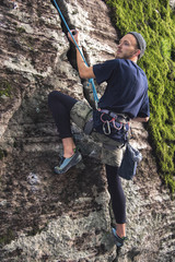 A young hipster is engaged in rock climbing with insurance on rocks with green moss