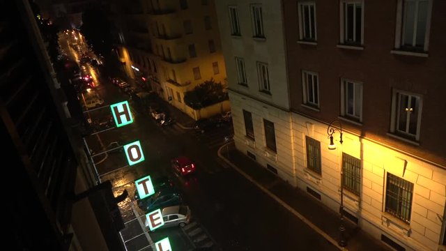 Footage from fourth floor looking down out of hotel window on streets during rainy night showing illuminated hotel sign and dark streets cars passing by with lights driving through evening darkness 4k