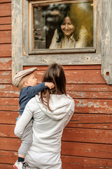 Rural scene with one year old baby boy with his mother outdoors looking at cottage window with grandmother inside