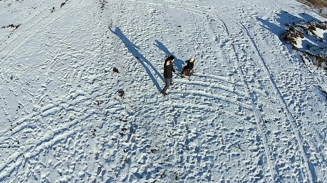 Aerial. Dad rolls his son on a sled and wave hands. Dady and baby have fun outdor. Man playing with boy and show like.