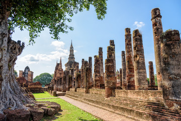 Ancient pagoda near a large tree in the ruins under the blue sky at Wat Maha That temple in Sukhothai Historical Park, old city and famous tourist attraction of Sukhothai Province, Thailand