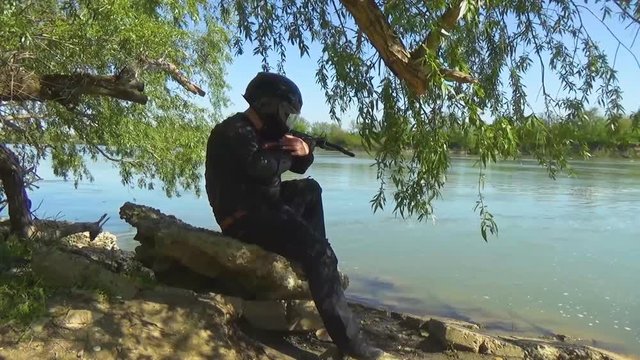 A Russian special forces soldier in the armor, closes the glass on his helmet and sits on a stone near the lake shore aiming from a crossbow. Marine recon.