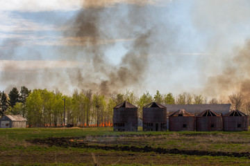 Fototapeta na wymiar A wall of fire with billowing smoke coming toward a line of green leaf trees and old abandoned wood grain bins in a agricultural summer landscape