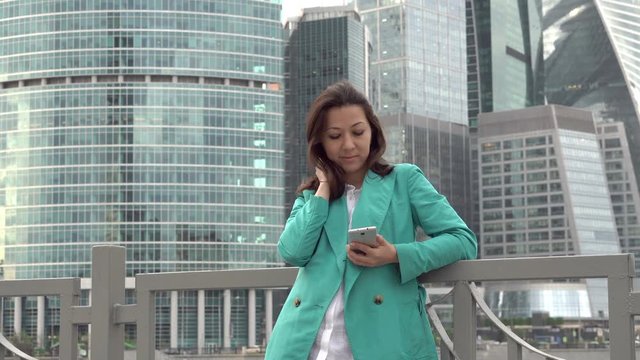 Stylish woman in a bright coat stands on the waterfront on the background of skyscrapers of business centers and use a smartphone, UHD 4K