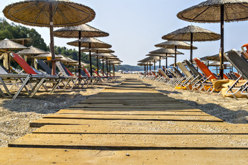 Wooden pathway on beautiful Ionian sandy resort beach in Greece with sunbeds and straw sunshades, umbrellas. Vacation destination. Public beach