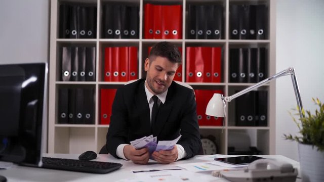 4k UHD. Young attractive businessman wearing a suit sitting in a desk counting euro bills in modern office