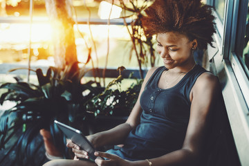 Smiling black curly teen sitting on the porch of resort summer house near pond and reading electronic book, cheerful afro american girl in t-shirt is making selfie on digital tablet near house window