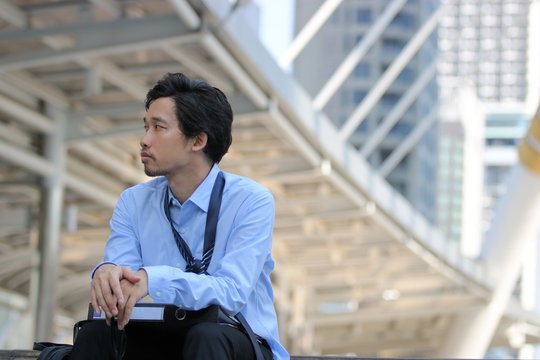 Frustrated stressed young Asian businessman sitting on the walkway and looking on far away in the city background.