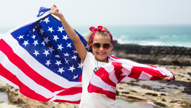 American flag. Little smiling patriotic girl holding an American flag flying in the wind on the ocean beach. National holiday July 4th, Independence day USA