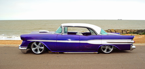 Classic  Purple American vintage car on seafront promenade, sea and beach in background. .