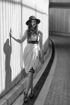 Young beautiful sexy woman wearing trendy outfit, white dress, black hat and leather swordbelt. Longhaired brunette posing in the city street. Outdoor fashion photography