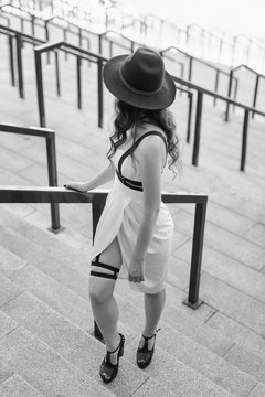 Young beautiful sexy woman wearing trendy outfit, white dress, black hat and leather swordbelt. Longhaired brunette posing in the city street. Outdoor fashion photography