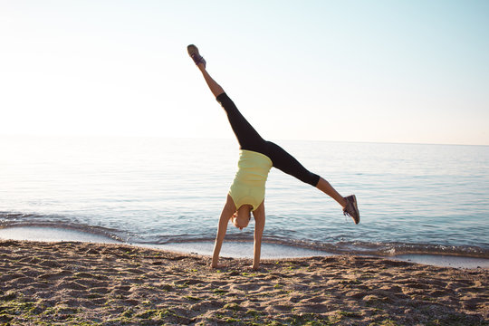 young professional gymnast woman dance on the beach,  training  exercises standing on hands, sunrise in sea or ocean background 