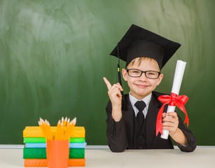 Happy boy with diploma in graduation hat points on empty green chalkboard