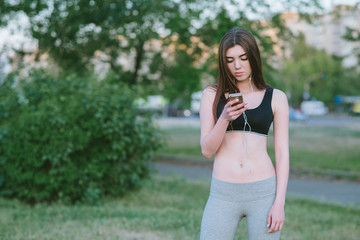 Fototapeta premium Athletic girl summer while jogging smartphone and enjoy listening to music in his headphones. Sports girl in a short shirt and leggings middle of the park with phone in hand.