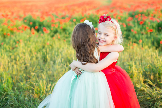 countryside, health, friendship, freedom, childhood, weekends concept - small fascinating fair-haired girl hugging her friend in light blue dress, they are sorrounded by poppies