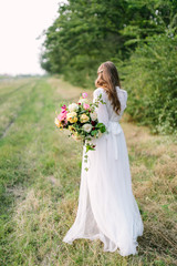 Fototapeta na wymiar wedding, marriage, beauty, nature, floral design, countrylife concept - tender silhouette of young bride in snowy white dress with flowing brown hair and great bouquet of peonies and soft roses