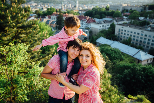 Portrait happy family of three. Father hugging mother and holding his son on the back. They standing and smiling on the hill with city view.