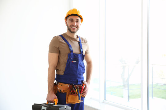 Young smiling electrician holding toolbox and standing near big window