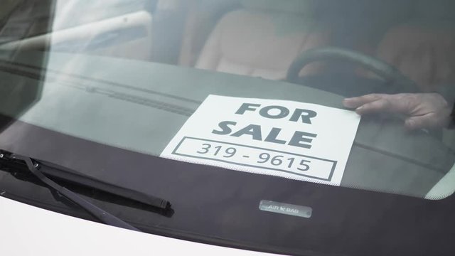Older man putting a FOR SALE sign on the dash of his vehicle.
