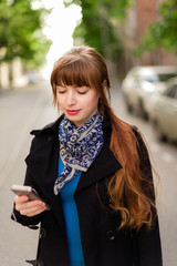 Beautiful red-haired young woman walking down the city street on a sunny day, using the Mobile Phone.