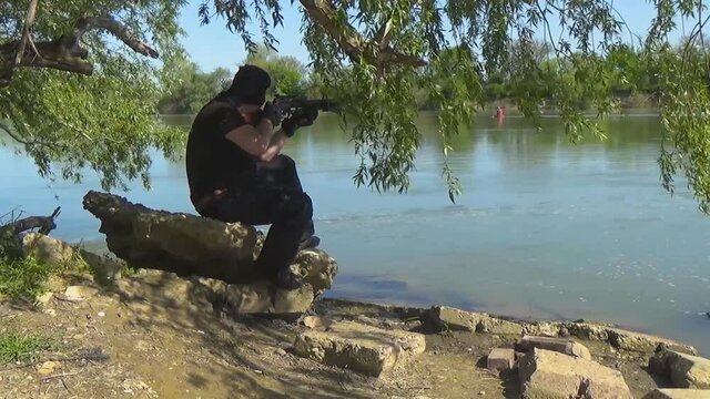 A Russian special forces soldier in a balaclava, sitting on a rock near the shore of the lake and aiming from a crossbow. Marine recon.