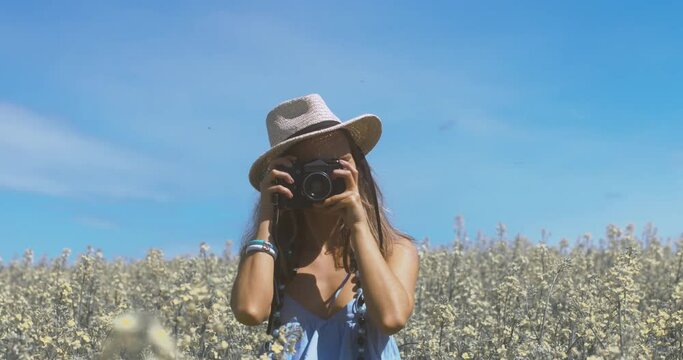 Attractive Caucasian female wearing blue dress and straw hat standing in a rapeseed field. Taking pictures with a vintage camera, then turning and running away from camera. 4K UHD RAW edited footage