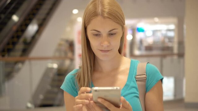 Young woman standing in shopping mall using smartphone, browsing, reading news, chatting with friends