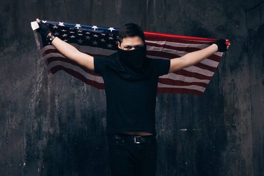 Flying USA flag and unrecognizable gangster. Guy in black cloth with hidden face on dark studio background. Riot, strike, bandit groups, disadvantaged youth concept